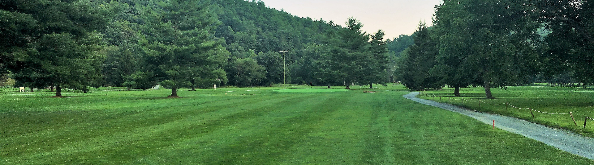 View of course hole with cart path on right side. A path of greens is visible in the middle with dark trees on both sides 